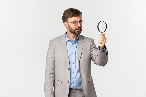 Портрет серьезного бизнесмена с бородой, в сером костюме и очках, ищущего что-то с увеличительным стеклом, исследующего на белом фоне — стоковое фото