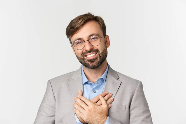 Närbild av stilig affärsman i grå kostym och glasögon, hålla händerna på hjärtat och tacka för något, ler tacksam, står över vit bakgrund — Stockfoto