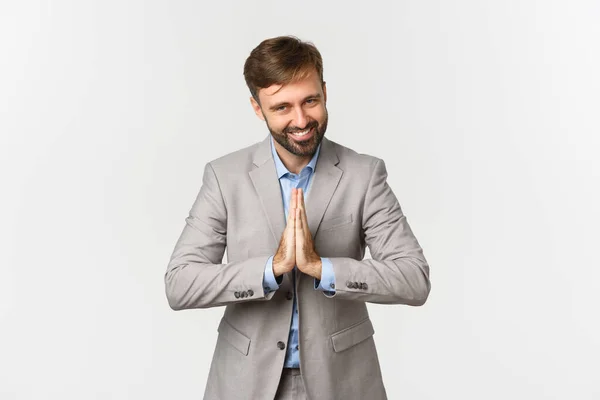 Portrait of happy bearded businessman in grey suit, thanking for something, holding hands pressed together and bowing with grateful expression, standing over white background — Stock Photo, Image