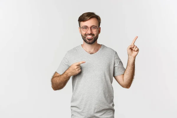 Homem barbudo alegre sorrindo, apontando os dedos no canto superior direito, mostrando logotipo, em pé sobre fundo branco — Fotografia de Stock