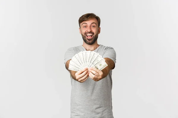 Imagen de tipo caucásico feliz con barba, dándole dinero y sonriendo, mostrando dinero en efectivo, de pie sobre fondo blanco — Foto de Stock