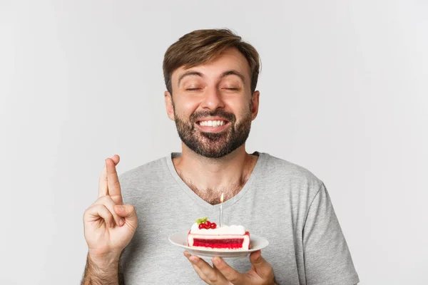 Close-up de homem esperançoso comemorando aniversário, fazendo desejo com os dedos cruzados, segurando bolo bday, de pé sobre fundo branco — Fotografia de Stock