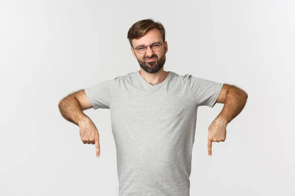 Disappointed bearded man grimacing, pointing fingers down, showing logo, wearing gray t-shirt, wearing gray t-shirt, standing over white background — Stock Photo, Image