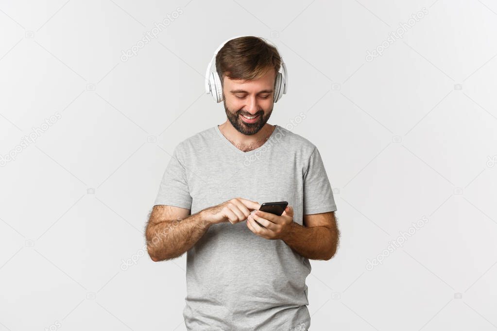 Portrait of attractive modern man smiling, looking at mobile phone, listening music in headphones, standing over white background