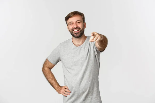 Image d'un caucasien insouciant avec barbe, portant un t-shirt gris, riant et pointant du doigt la caméra, debout sur fond blanc — Photo