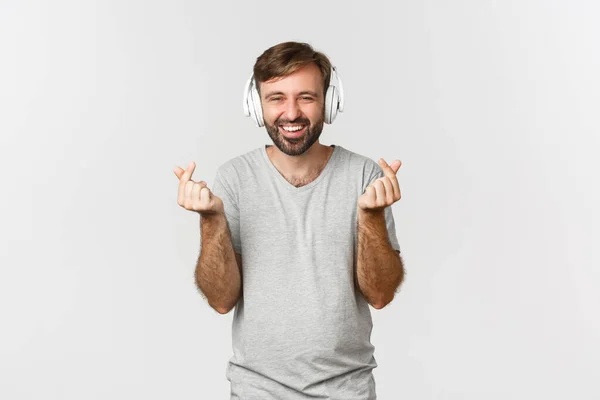 Beeld van vrolijke blanke man in grijs t-shirt, luisterend naar muziek in hoofdtelefoon en hartklachten, glimlachend op camera, staand over witte achtergrond — Stockfoto