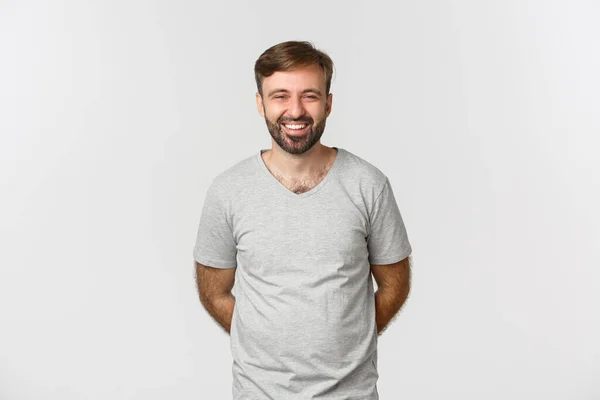 Imagen de hombre barbudo feliz en camiseta gris, sonriendo y riendo, de pie humilde con las manos detrás — Foto de Stock