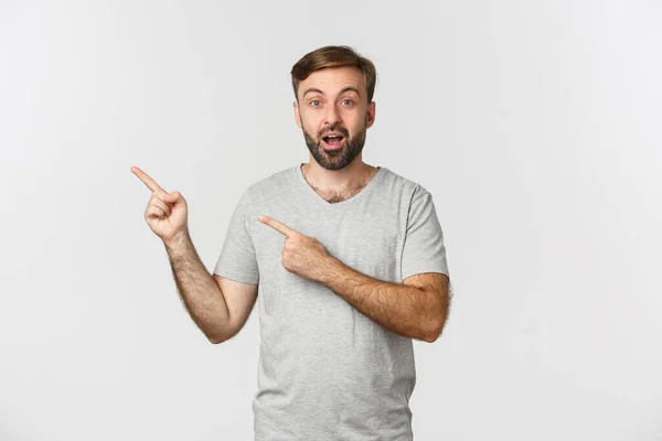 Bild av förvånad skäggig kille i grå t-shirt, pekar finger på övre vänstra hörnet logotyp, visar reklam, står över vit bakgrund — Stockfoto