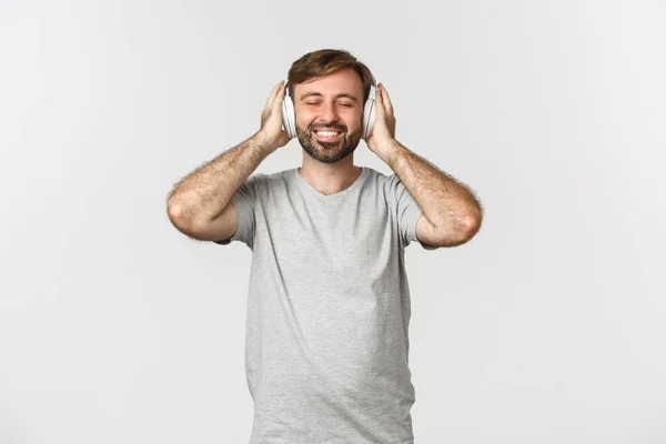Bild av stilig man i casual outfit, lyssna musik i trådlösa hörlurar, leende nöjd, står över vit bakgrund — Stockfoto