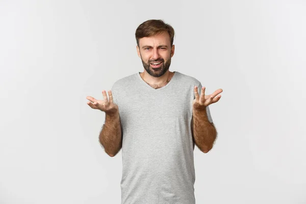 Porträtt av besviken och förvirrad man med skägg, lyfta händerna upp och klaga, står i grå t-shirt över vit bakgrund — Stockfoto