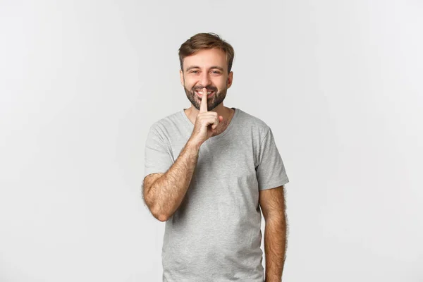 Retrato de belo homem sorridente em camiseta cinza, calado com você gentilmente pedindo para ficar quieto, em pé sobre fundo branco — Fotografia de Stock