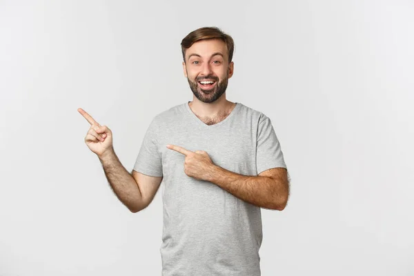 Muotokuva innoissaan hymyilevä mies parta, yllään rento harmaa t-paita, sormet vasemmassa yläkulmassa, osoittaa logo, seisoo valkoisen taustan — kuvapankkivalokuva