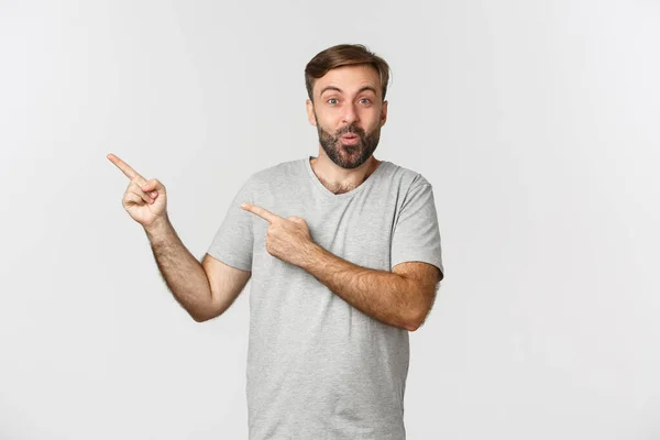 Retrato de homem barbudo animado e surpreso em camiseta cinza, apontando os dedos no canto superior esquerdo, mostrando o logotipo, em pé sobre o fundo branco — Fotografia de Stock