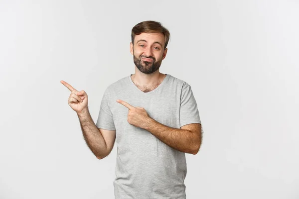 Portret van een sceptische man met een baard in een grijs t-shirt, met iets stoms erop, wijzend met vingers naar de linkerbovenhoek en kijkend naar een ongeamuseerde, witte achtergrond — Stockfoto