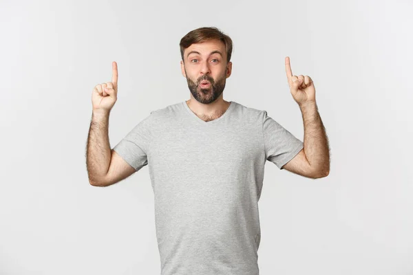 Imagem de homem bonito animado em camiseta cinza, apontando os dedos para cima e dizendo uau, de pé sobre fundo branco — Fotografia de Stock