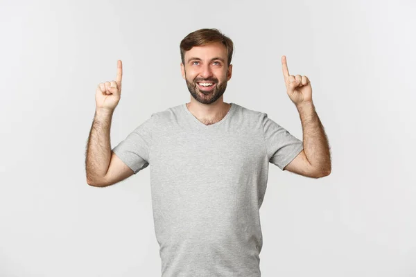 Retrato de um cara sorridente com uma barba, vestindo camiseta cinza, apontando os dedos para o logotipo, em pé sobre o fundo branco — Fotografia de Stock