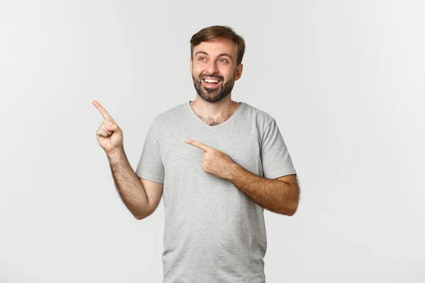 Bonito homem sorridente em camiseta cinza, olhando sonhador no canto superior esquerdo, mostrando logotipo, de pé sobre fundo branco — Fotografia de Stock