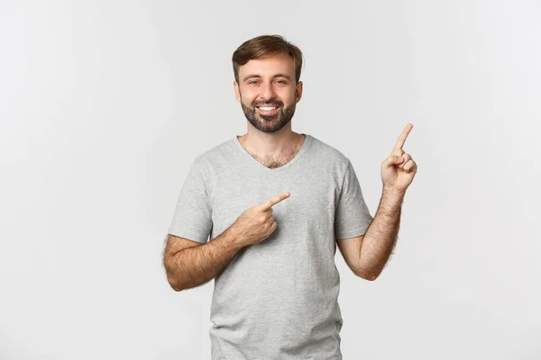 Imagen de un chico guapo feliz con barba, vistiendo una camiseta gris, señalando con los dedos el logo de la esquina superior derecha y sonriendo, de pie sobre fondo blanco —  Fotos de Stock