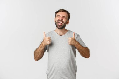 Sakallı mutlu ve heyecanlı bir adamın portresi, komik bir şeye gülüyor ve baş parmağını kaldırıyor, beyaz arka planda duruyor.
