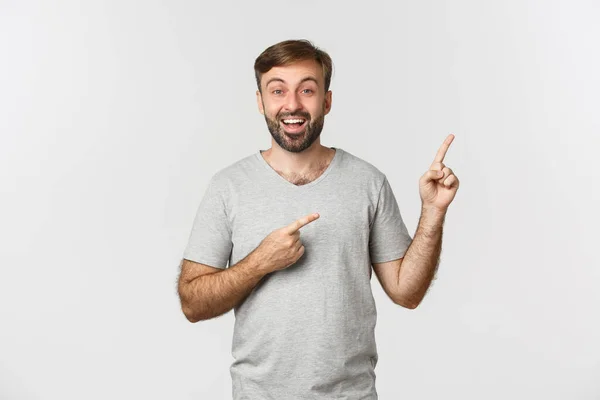 Feliz chico adulto guapo con barba, usando camiseta, señalando los dedos en la esquina superior derecha, mostrando pancarta, de pie sobre fondo blanco — Foto de Stock