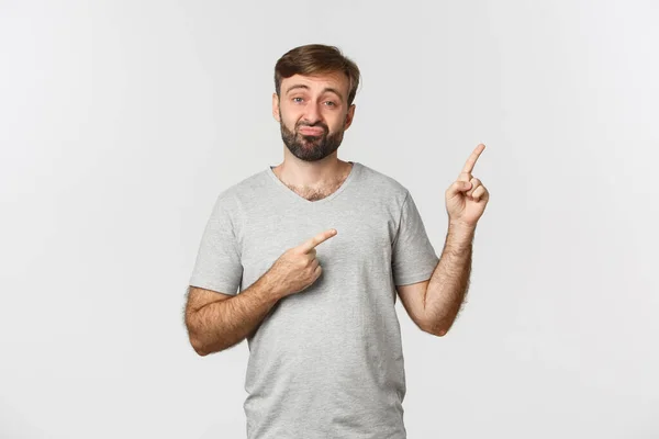 Skeptical and disappointed guy with beard, sulking and pointing at upper right corner, showing lame and unimpressive product, standing over white background — Stock Photo, Image