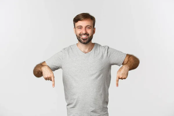 Retrato de homem barbudo atraente em camiseta cinza, mostrando logotipo, apontando os dedos para baixo, em pé sobre fundo branco — Fotografia de Stock