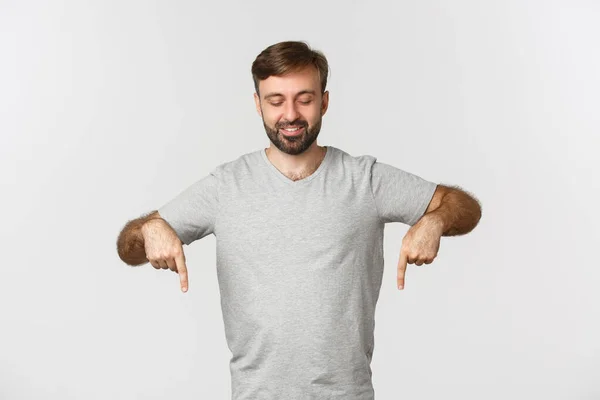 Attraente modello maschio caucasico in t-shirt grigia, punta e guarda in basso, sorridente soddisfatto, in piedi su sfondo bianco — Foto Stock