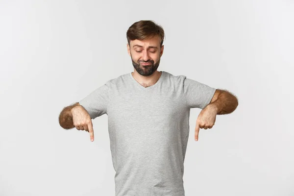 Porträt eines skeptischen bärtigen Mannes im grauen T-Shirt, der die Stirn runzelt und mit den Fingern nach unten zeigt, etwas Schlechtes zeigt, vor weißem Hintergrund steht — Stockfoto