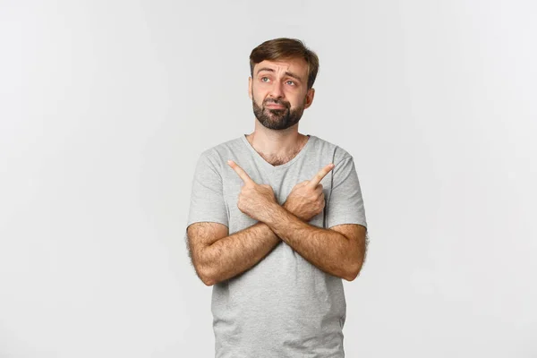 Portrait of indecisive beaded man in gray t-shirt, making decision, pointing fingers sideways at two choices, standing over white background — Stock Photo, Image