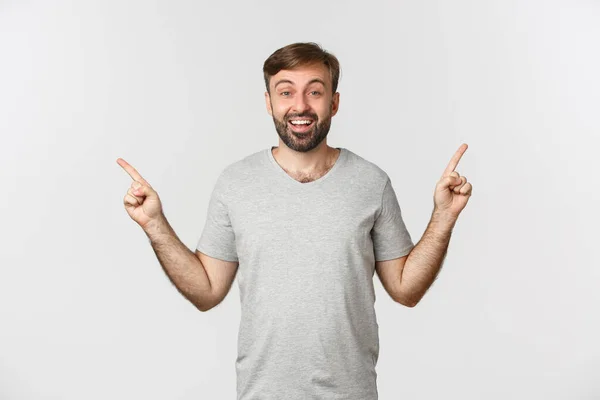 Porträtt av glad leende man med skägg i grå t-shirt, pekar finger i sidled, visar två bra val, står över vit bakgrund — Stockfoto