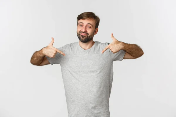 Portrait d'un bel homme barbu en t-shirt gris pointant le logo et souriant, recommandant la marque, debout sur fond blanc — Photo