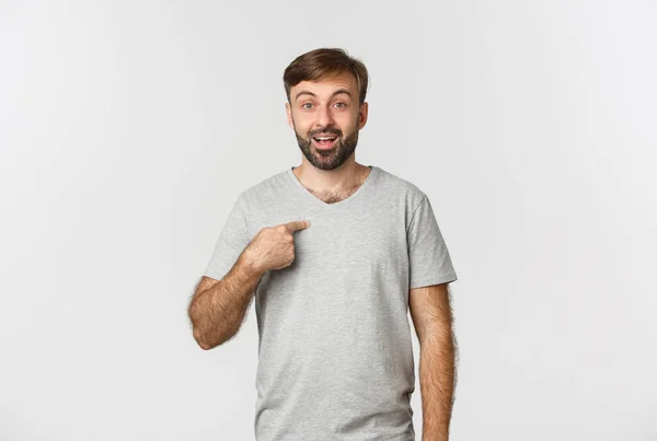 Imagen del hombre feliz y sorprendido en camiseta gris, apuntándose a sí mismo, de pie sobre fondo blanco — Foto de Stock