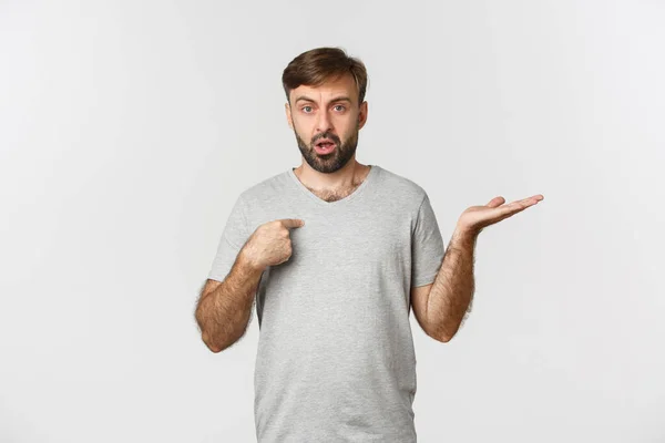 Retrato de hombre barbudo guapo en camiseta gris, apuntándose a sí mismo y encogiéndose de hombros confundido, siendo acusado, de pie sobre fondo blanco —  Fotos de Stock