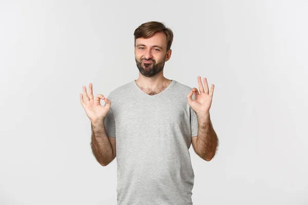 Portrét spokojený pohledný chlap s vousy, ukazuje v pořádku znamení a schválit něco dobrého, chvályhodný výběr, stojící nad bílým pozadím — Stock fotografie