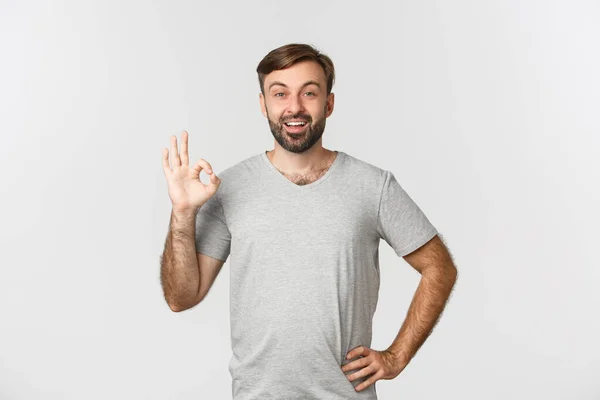 Portret van tevreden knappe man met baard, tonen oke teken en goedkeuren iets goeds, prijzenswaardige keuze, staande over witte achtergrond — Stockfoto