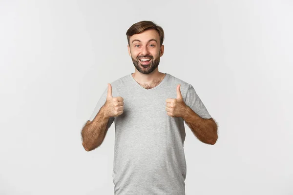 Imagem de homem barbudo bonito em camiseta cinza, parecendo animado, mostrando polegares para cima na aprovação, como algo bom, de pé sobre fundo branco — Fotografia de Stock