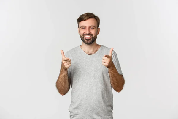 Imagem de homem sorridente satisfeito em camiseta cinza, mostrando polegares para cima na aprovação, como algo bom, fundo branco — Fotografia de Stock