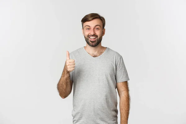 Kuva tyytyväinen hymyilevä mies harmaa t-paita, osoittaa peukalot ylöspäin hyväksyntää, kehua hyvä valinta, valkoinen tausta — kuvapankkivalokuva