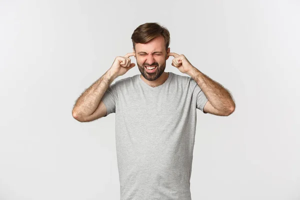 Porträtt av irriterad kille i grå t-shirt, stänga öronen och grimma från högt ljud, står över vit bakgrund — Stockfoto