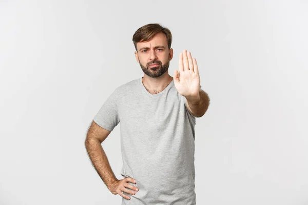 Porträtt av allvarliga rynka pannan kille i grå t-shirt, visar stopp gest, ogillar och förbjuda åtgärder, står över vit bakgrund — Stockfoto