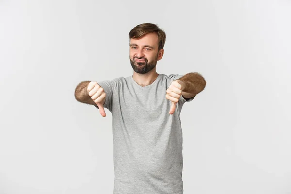 Portrait of skeptical bearded man in casual t-shirt, showing thumbs-down and looking disappointed, disapprove something bad, standing over white background — Stock Photo, Image