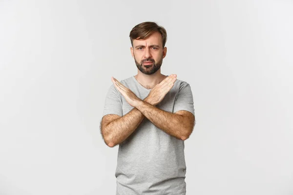 Разочарованный человек с бородой, показывающий крестовый жест и хмурящийся, говорящий остановиться, запрещающий что-то плохое, стоящий на белом фоне — стоковое фото