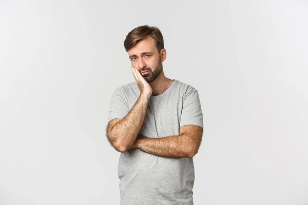 Retrato de homem barbudo triste e entediado em camiseta cinza, inclinado na palma da mão e olhando relutante, em pé sobre fundo branco — Fotografia de Stock