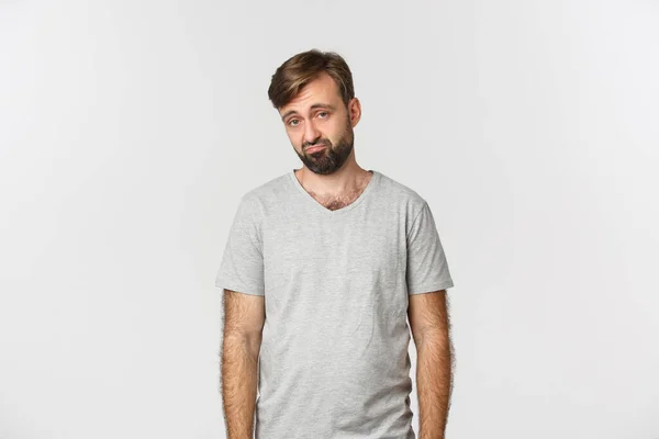 Muotokuva surullinen ja synkkä parrakas mies harmaa t-paita, murjottaa ja näyttää kurja, seisoo valkoisella taustalla — kuvapankkivalokuva