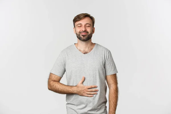 Muotokuva onnellinen mies tunne tyytyväinen syötyään maukasta ruokaa, hankausta vatsa ja hymyillen iloinen, seisoo valkoisella taustalla — kuvapankkivalokuva