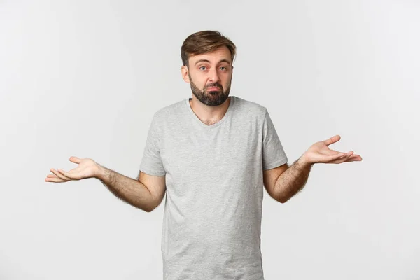 Imagem do homem barbudo sem noção em t-shirt casual, encolhendo os ombros e olhando confuso, de pé sobre fundo branco — Fotografia de Stock