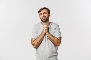 Gri tişörtlü yakışıklı bir adamın portresi, bir şey için yalvarıyor, yalvarıyor ve perişan görünüyor, beyaz arka planda duruyor.