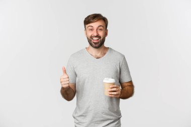 Gri tişörtlü neşeli beyaz adam portresi, kahve içiyor ve baş parmağını kaldırıyor, tavsiye ediyor ve kafe gibi beyaz arka planda duruyor.