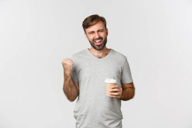 Sakallı yakışıklı bir adamın portresi, gri temel tişört giyiyor, kahve içtikten sonra enerji dolu hissediyor, yumruk pompası yapıyor ve heyecanla evet diyor, beyaz arka planda duruyor.