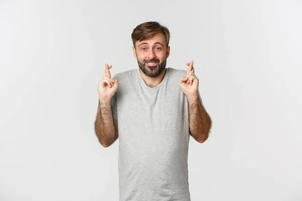 Portrait of hopeful smiling man with beard, wearing gray t-shirt, crossing fingers for good luck and looking at camera, making wish, standing over white background — Stock Photo, Image